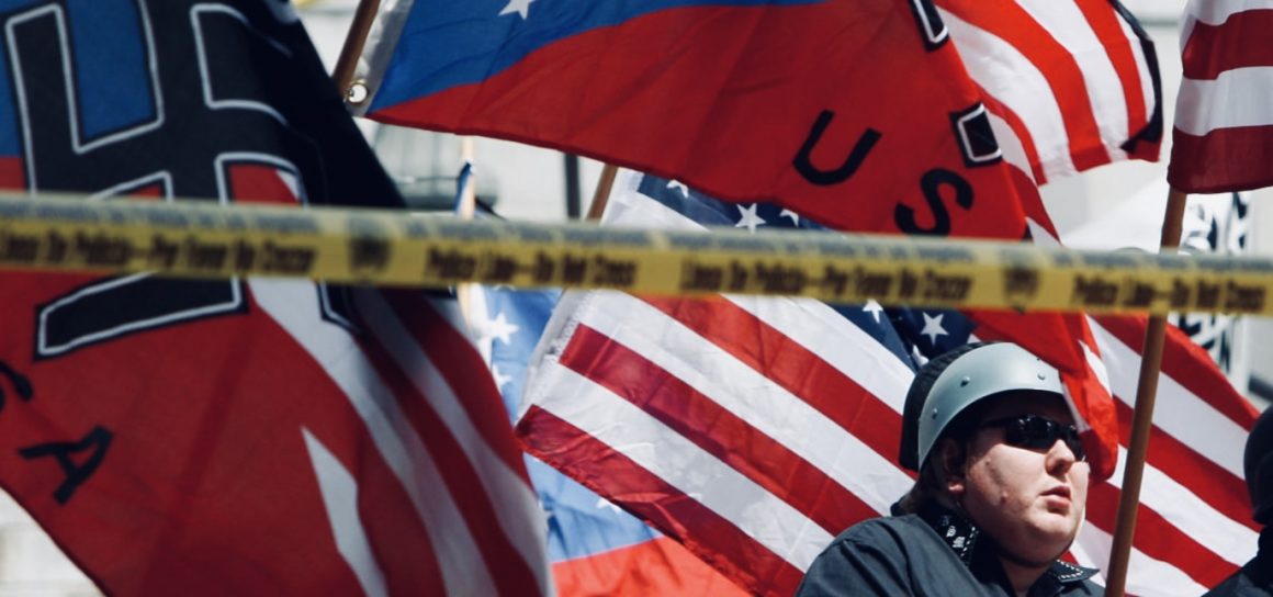 An image showing a police 'do not cross' line and behind it is a man wearing sun glass and a helmet and behind him are a number of flags with American and Nazi symbols.